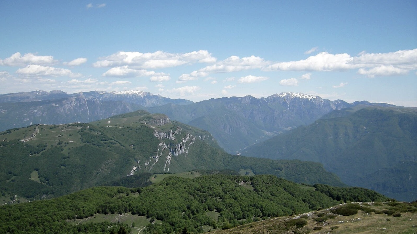 A scenic landscape of the Italian Alps showcasing a range of mountains with varying elevations. The foreground features rolling green hills with sparse tree coverage, leading to rugged mountain peaks in the distance